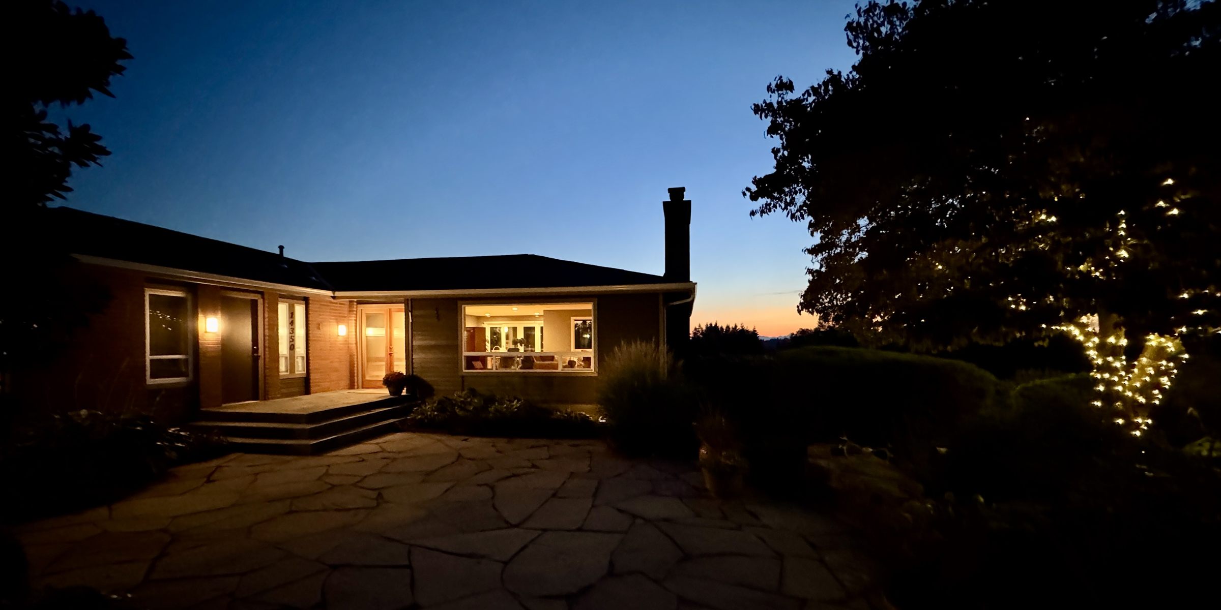 The house at dusk: warm light in the big front window, a flagstone courtyard, and string lights in the trees against a blue and orange sky.